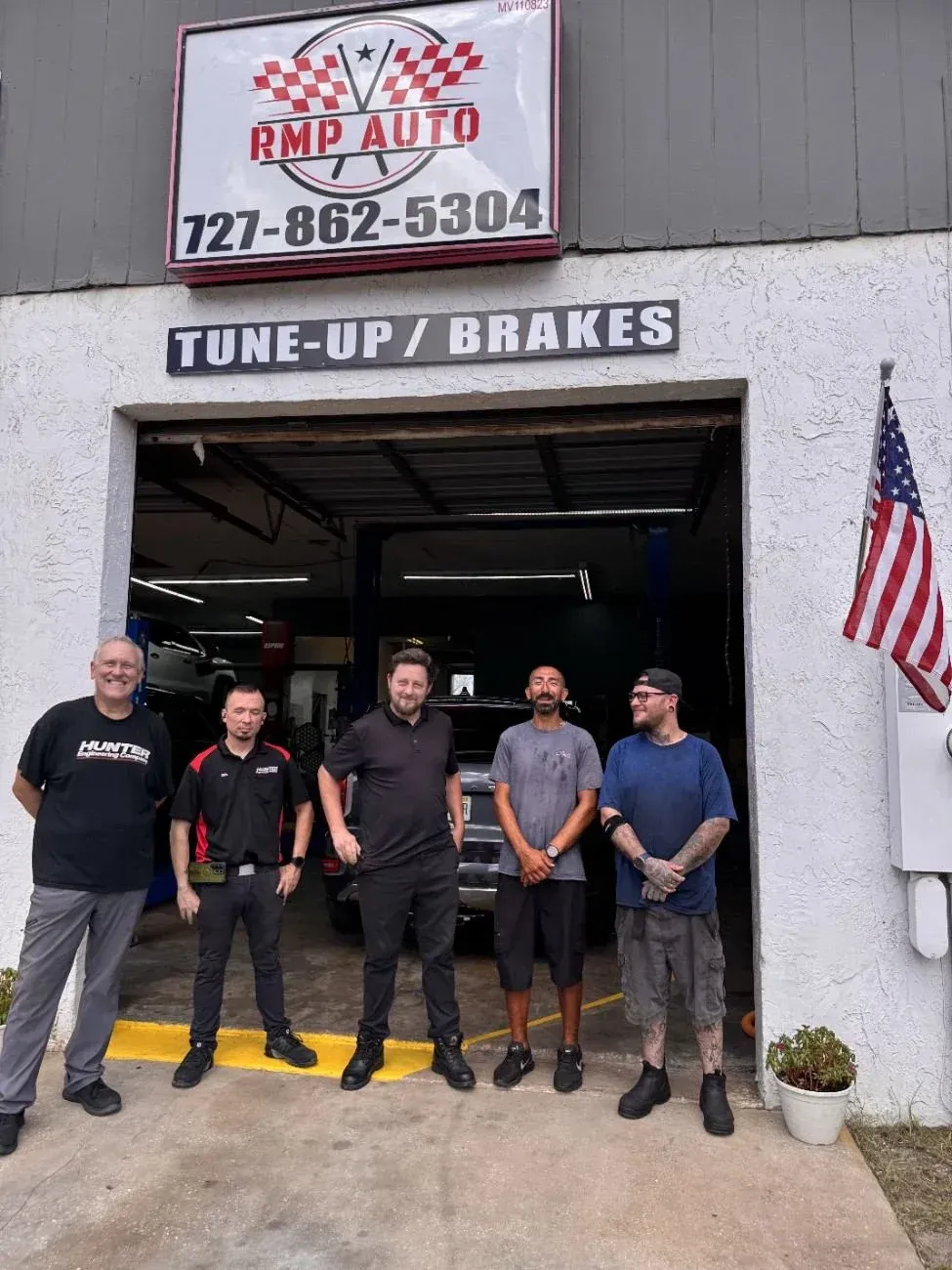 Five mechanics standing outside RMP Auto garage under a TUNE-UP/BRKES sign and American flag