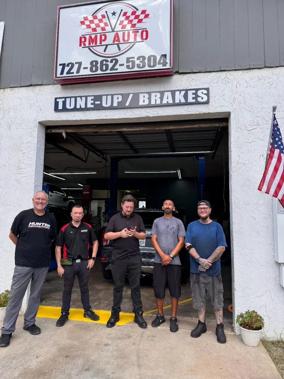Five men standing outside RMP Auto garage under a “TUNE-UP/ BRAKES” sign, with an American flag beside the door