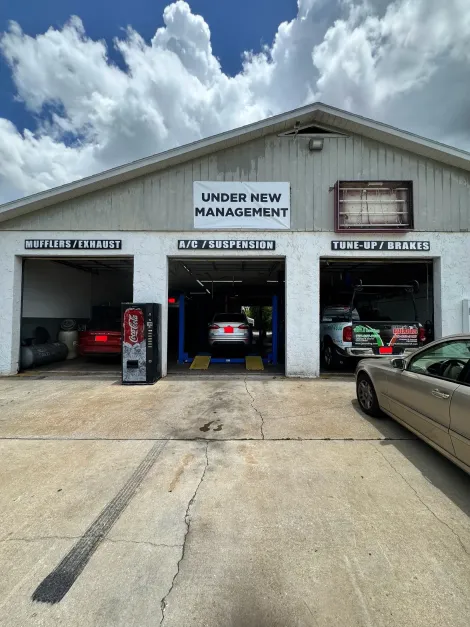 Car repair shop with three open service bays and cars inside, under a cloudy sky, one car in the foreground