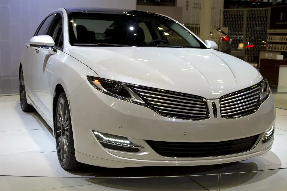 White sedan displayed indoors on a showroom floor, front view with chrome grille and headlights.