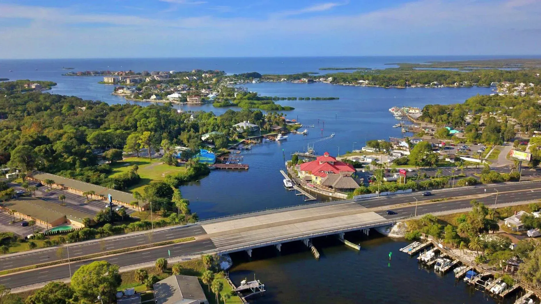 Aerial view of a coastal town with a river, bridge, boats, and houses near the water under a blue sky