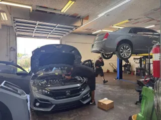 Auto repair shop with two cars, one hood open and another raised on a lift, mechanic nearby.