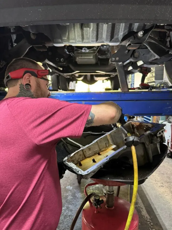 Mechanic in pink shirt works under a raised vehicle, handling a metal engine pan on a red stand.