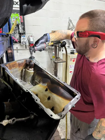 Mechanic wearing red goggles inspects an open car engine compartment in a garage