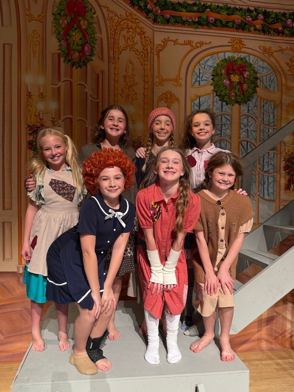 Seven smiling children in colorful costumes posing on a stage set with painted floral backdrop.