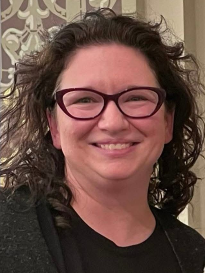 Smiling person with curly hair and glasses, wearing a black top against a patterned background