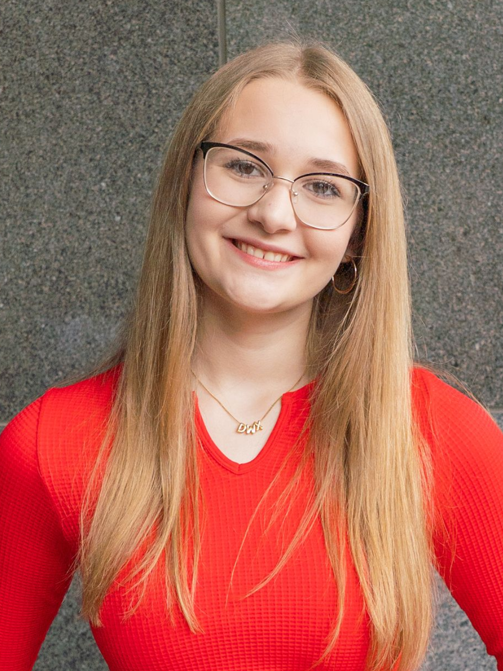 Smiling person with long blonde hair and glasses wearing a red top against a gray wall