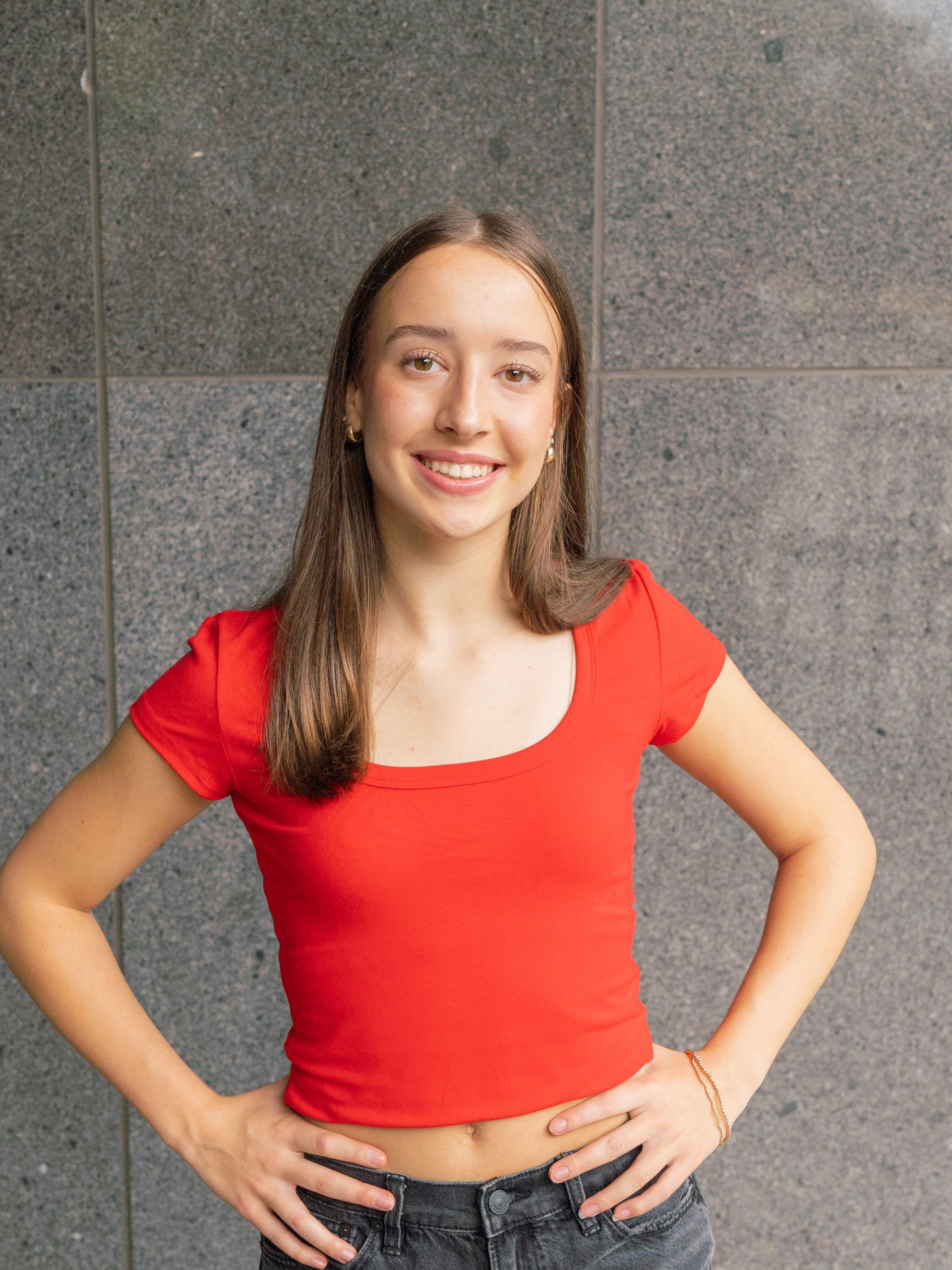 Smiling person in a red top and jeans, standing with hands on hips against a gray wall.