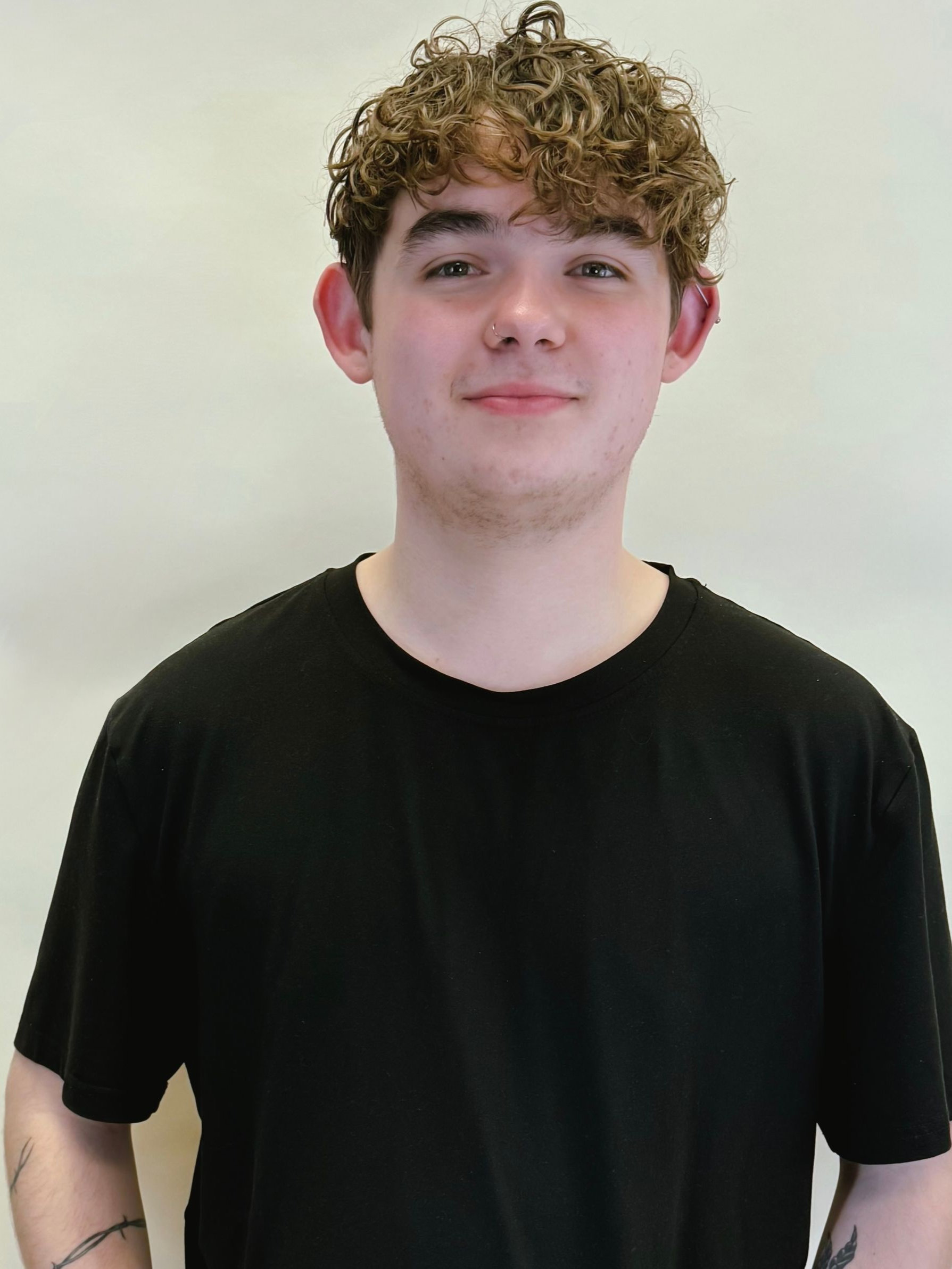 Person with curly hair wearing a black T-shirt, standing against a plain light background.