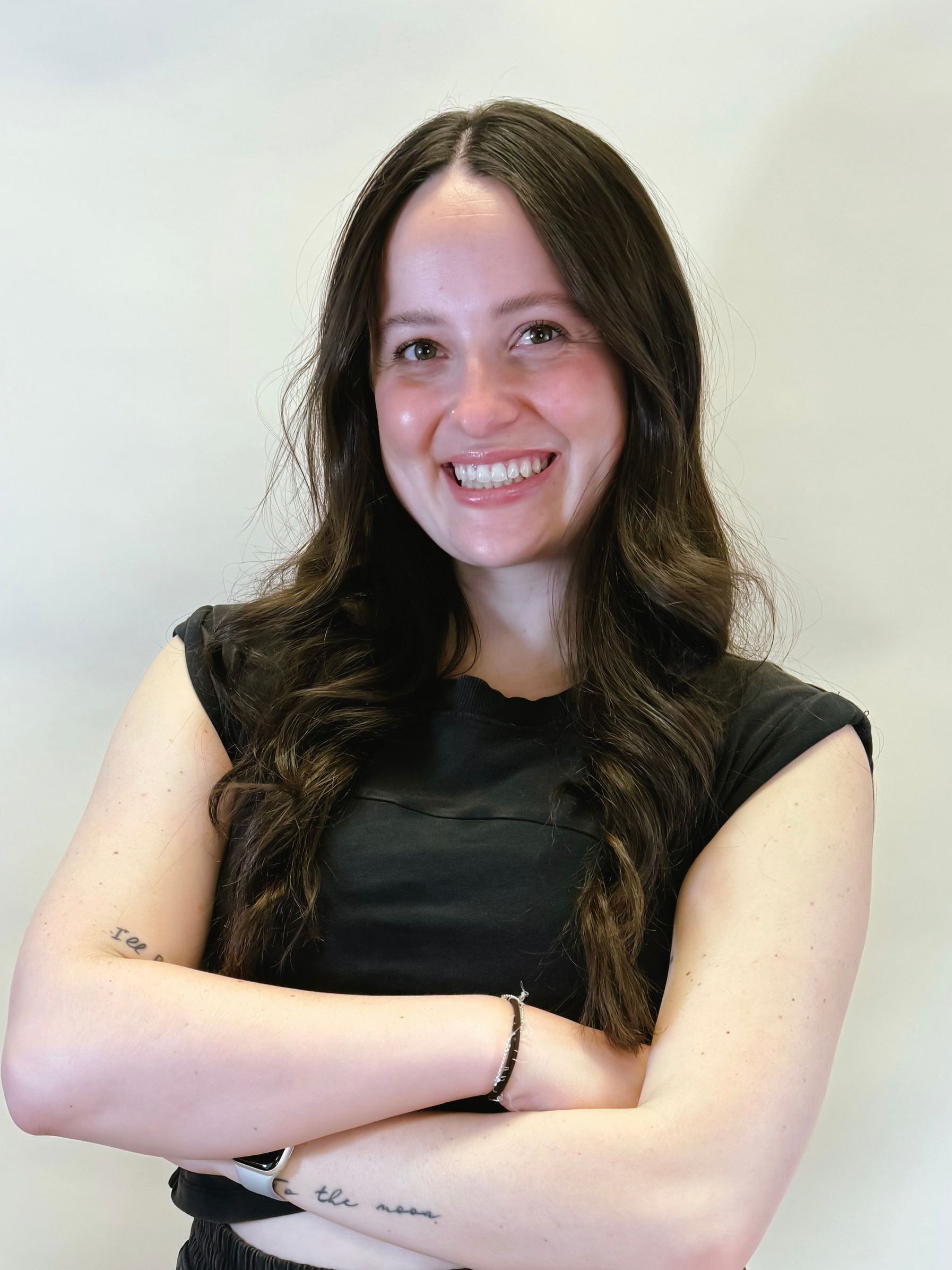 Smiling person with long dark hair, wearing a black top and arms crossed against a plain light background
