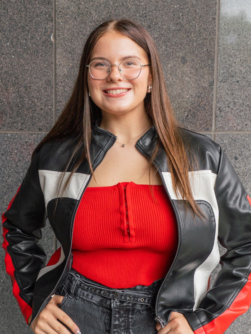 Smiling person in glasses, red top, and black-and-silver jacket standing against a gray stone wall.