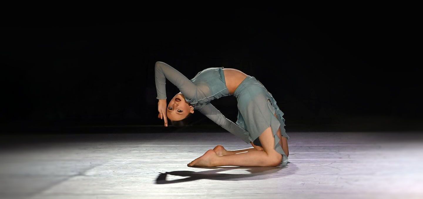 Contemporary dancer in a gray outfit performing a deep backbend on a stage under a spotlight
