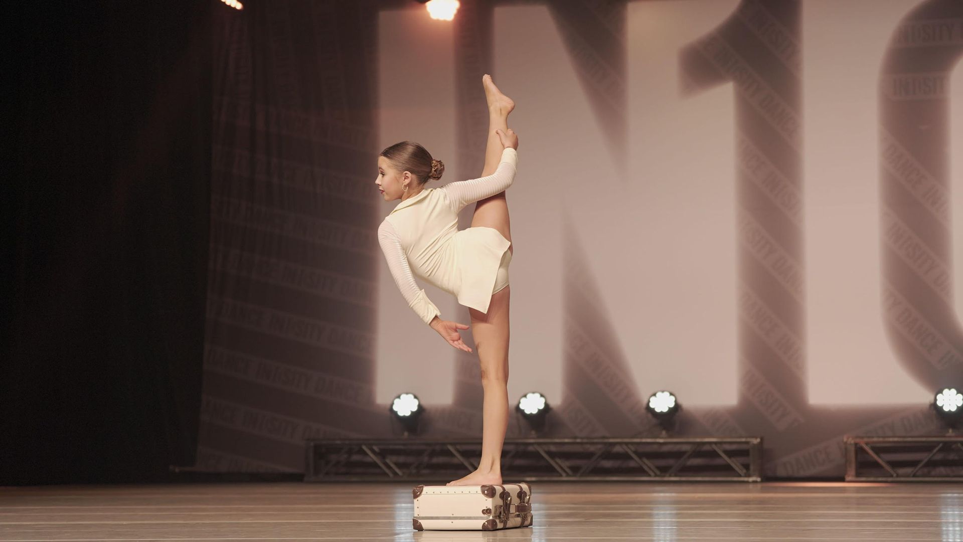 Dancer in white costume balancing on one leg with leg lifted high on a lit stage