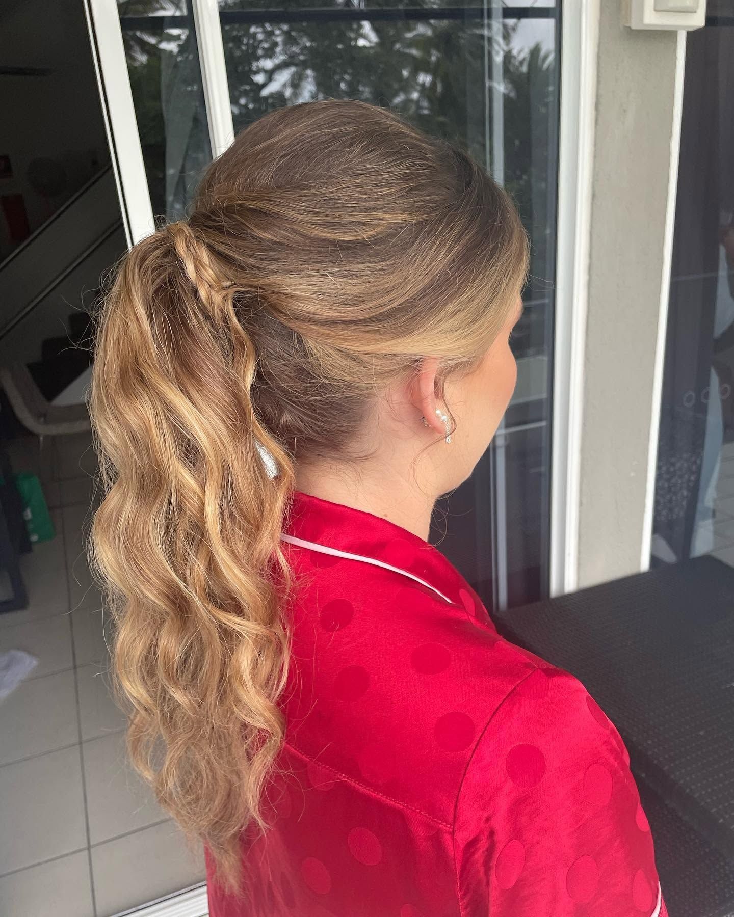 Woman with blonde wavy ponytail wearing a red shirt, seen from behind near a doorway.