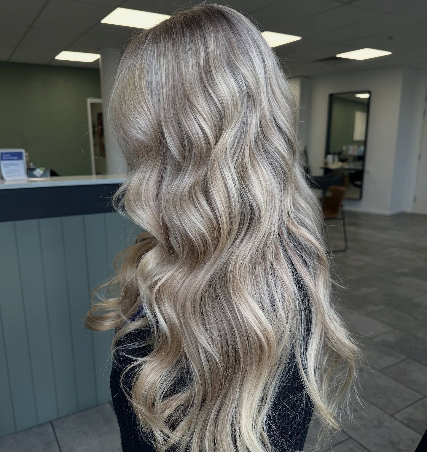 Wavy platinum-blonde hair styled in loose curls, viewed from behind in a salon.