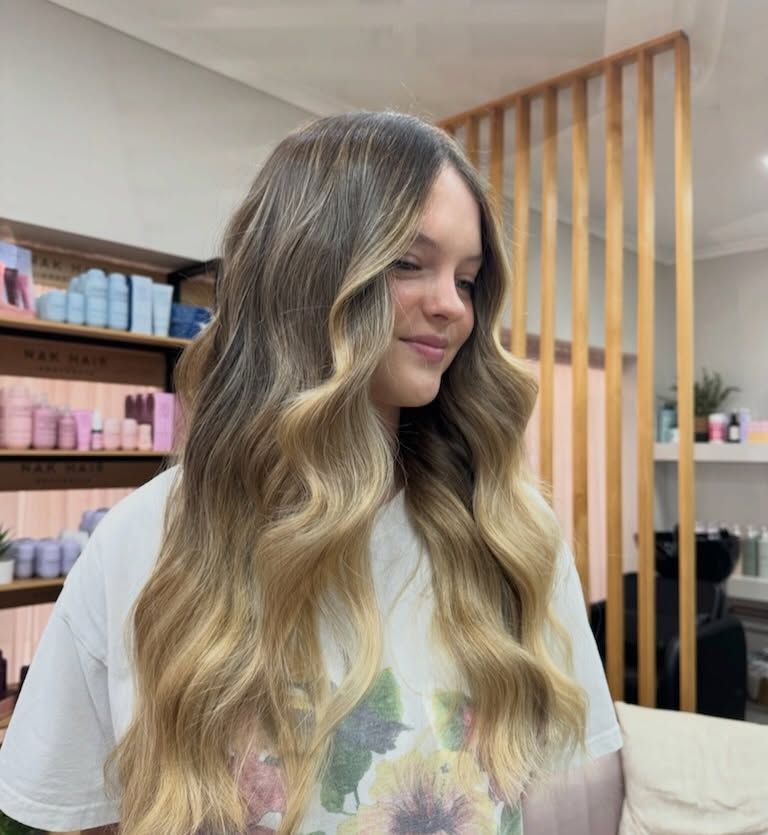 Woman with long wavy blonde hair smiling in a salon, with shelves of hair products in the background — Blackwell Hair In Woree, QLD