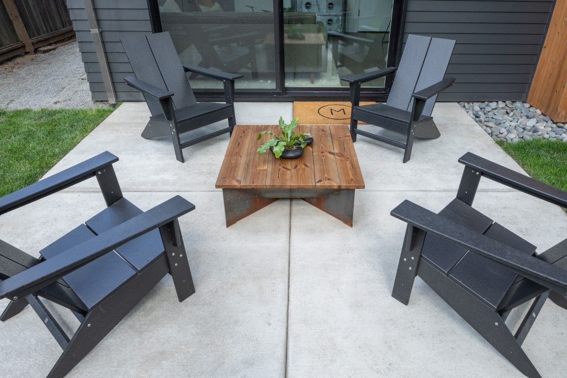 Four black Adirondack chairs surrounding a square wooden coffee table on a concrete patio.