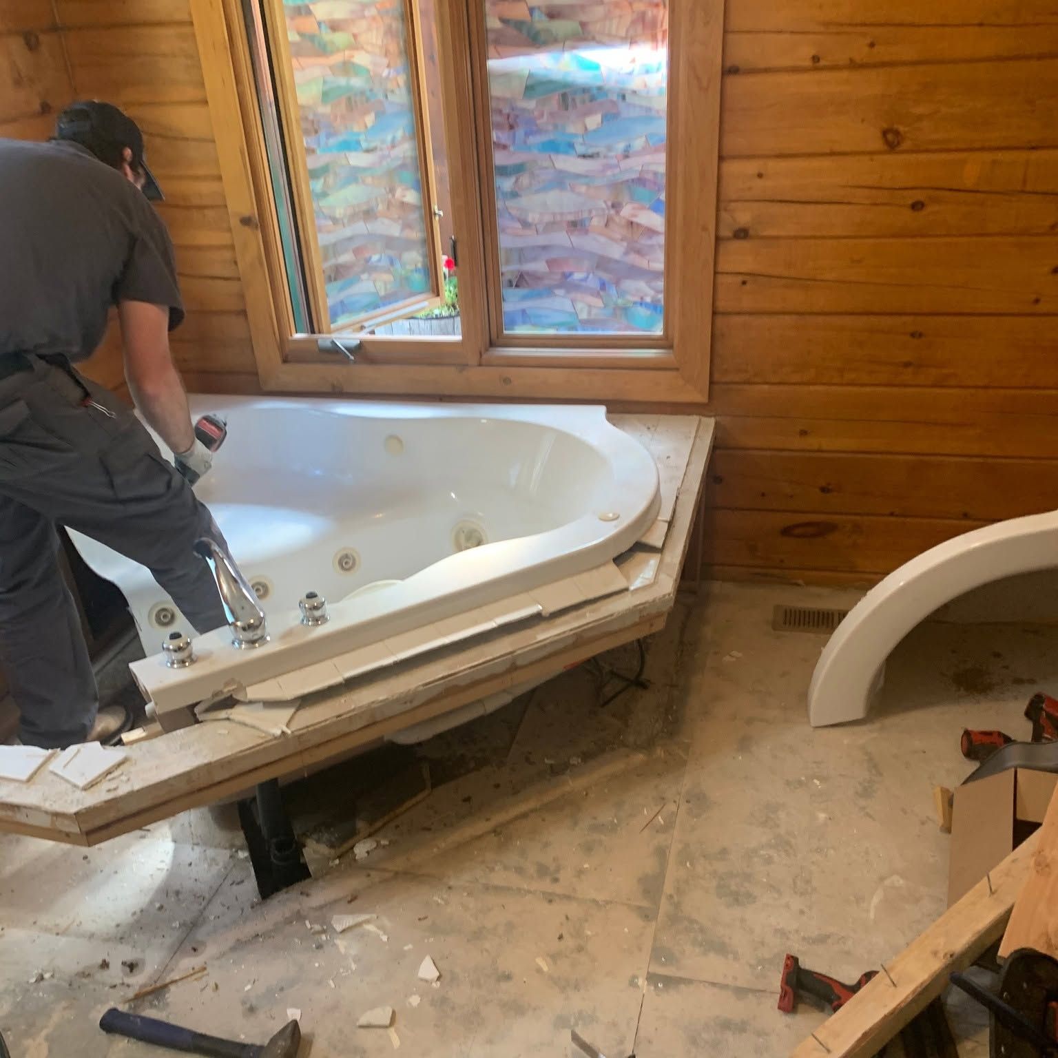 Worker installing a white bathtub in a wood-paneled bathroom with a window and construction debris.