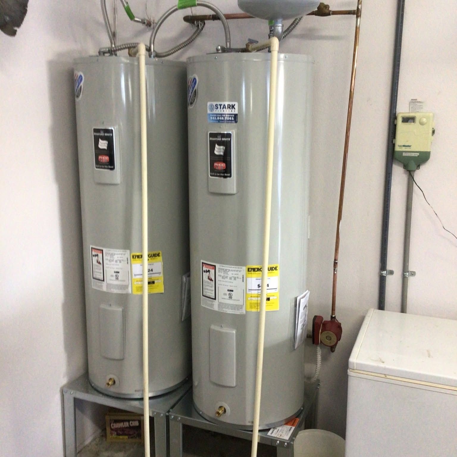 Two tall silver water heaters in a utility room, with pipes and a white appliance beside them.