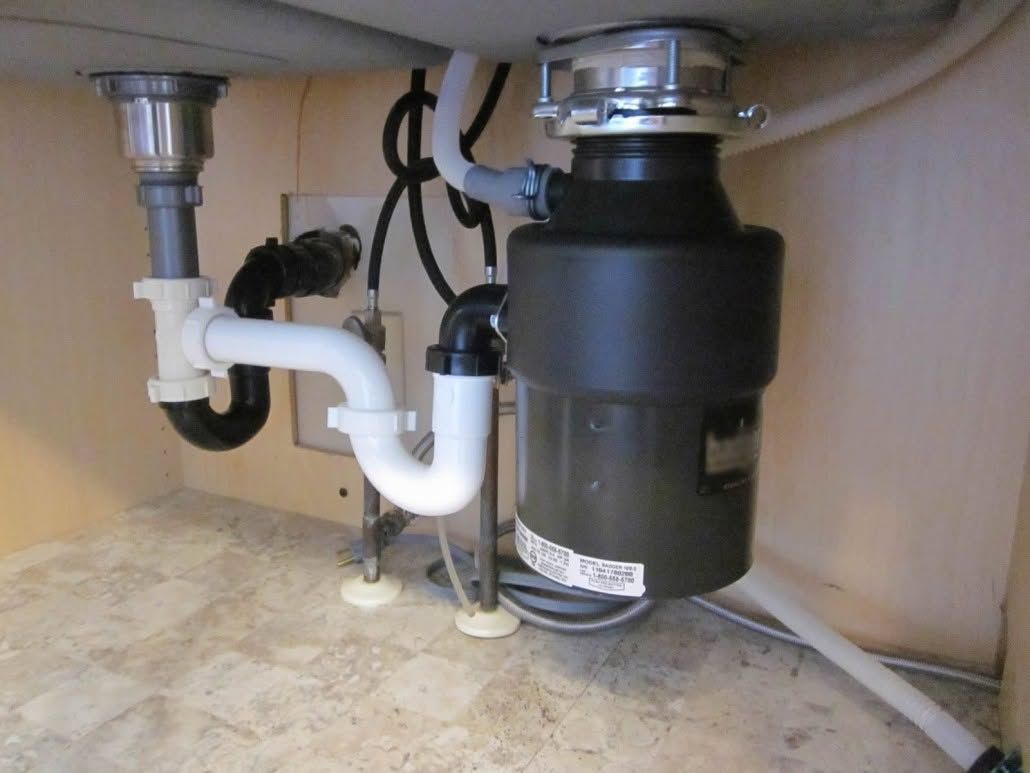 Under-sink plumbing with white drain pipes and a black garbage disposal unit.