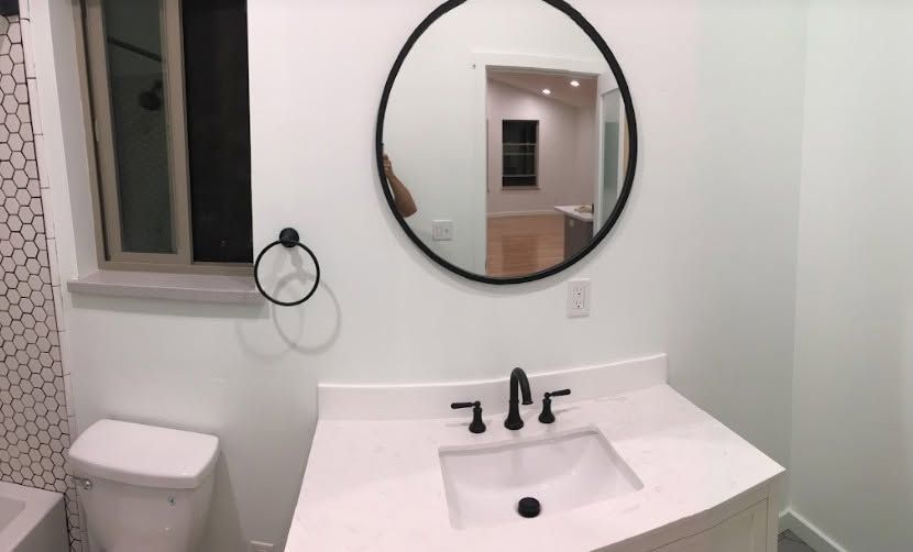 Modern bathroom vanity with round mirror, white countertop, black fixtures, and toilet by a window