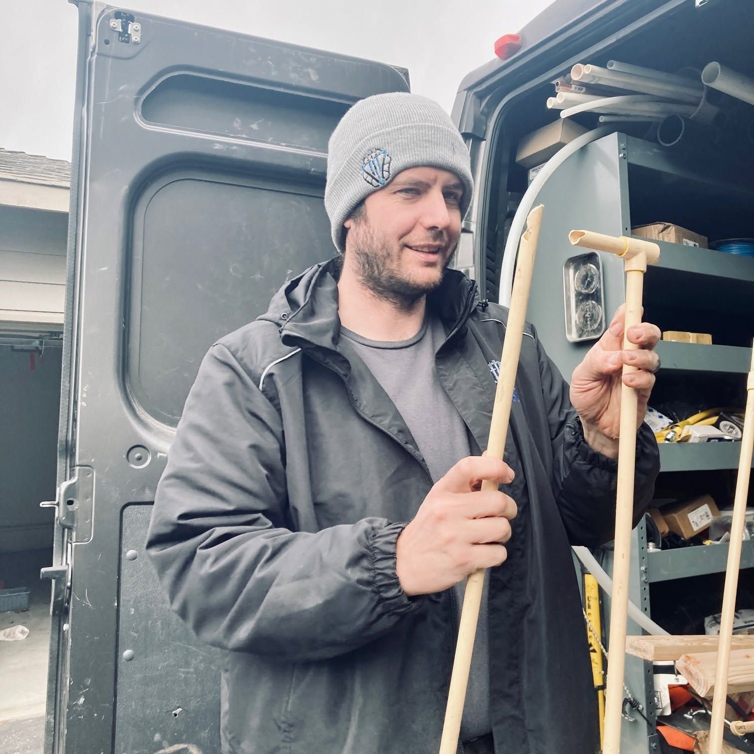 Man in gray beanie and jacket holding a wooden tool beside an open van with shelves of supplies