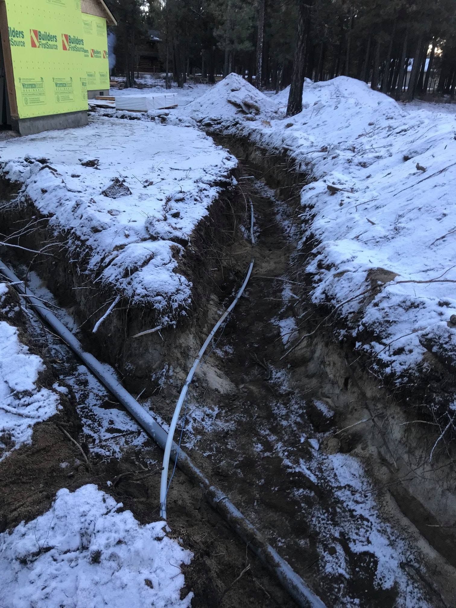 Snowy muddy trench with hoses running through a wooded area beside a yellow building