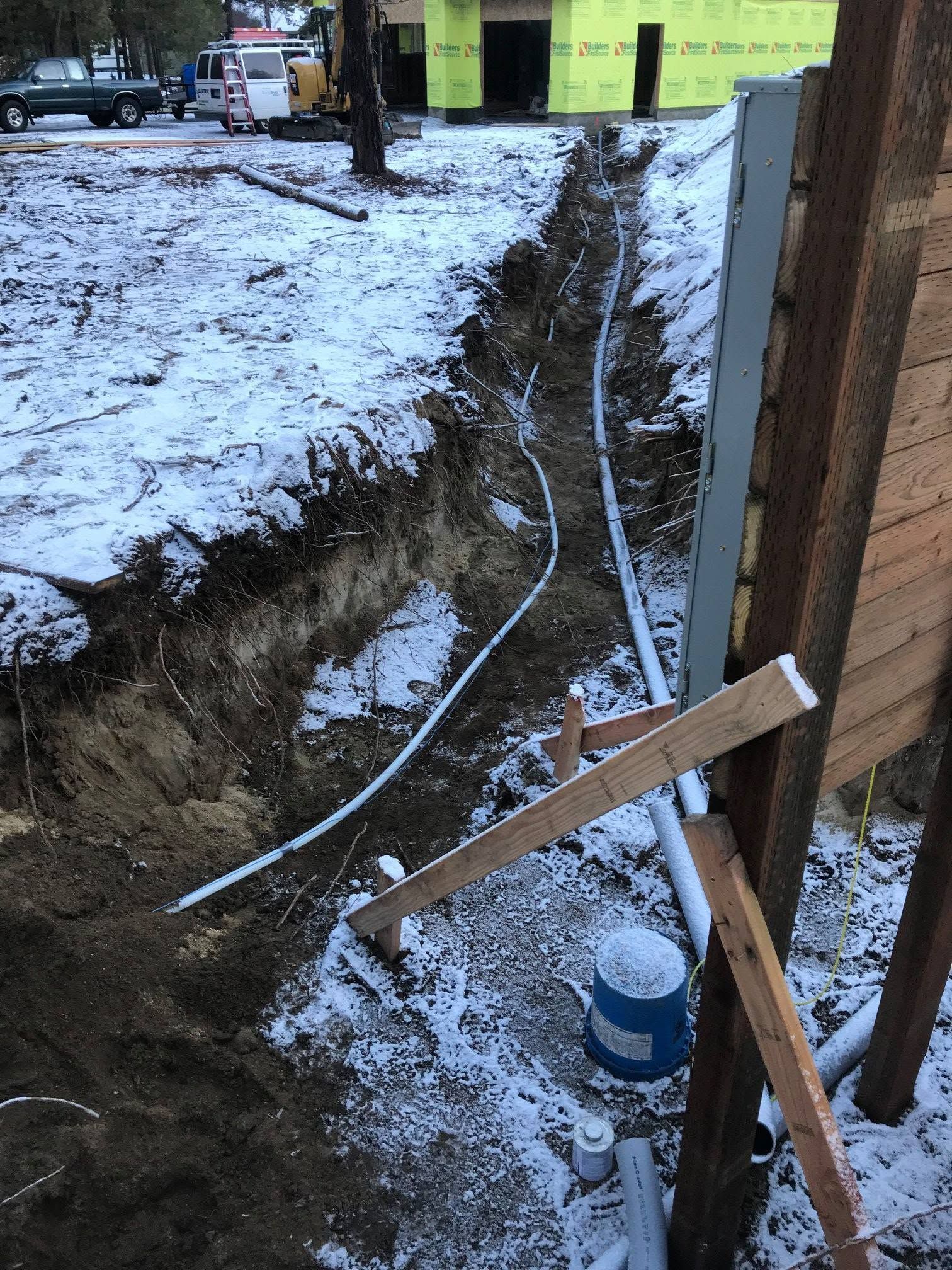Snowy trench with exposed pipes and a wooden brace beside a brick wall.
