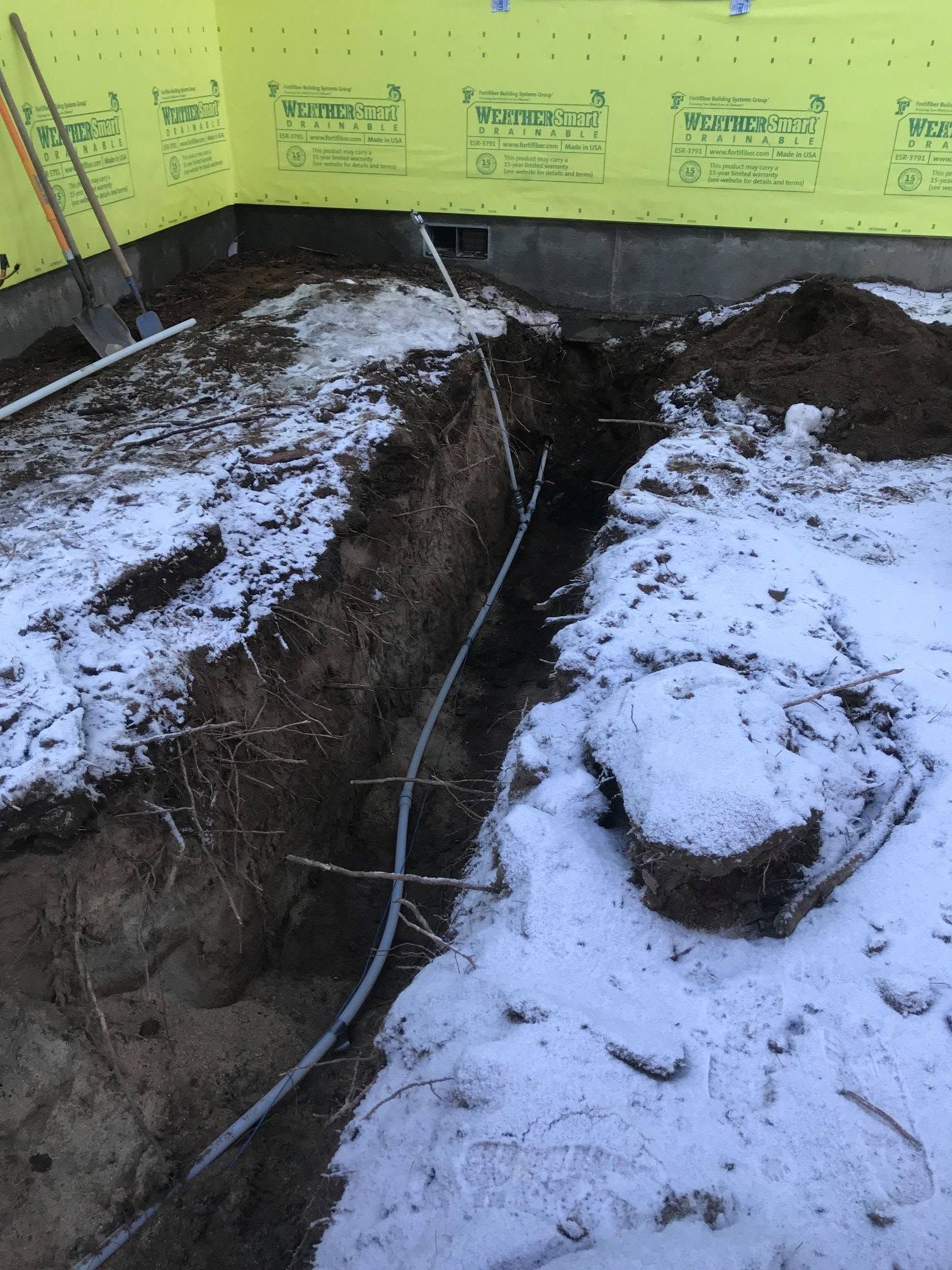 Snow-covered excavation trench beside a yellow foundation wall, with exposed dirt and a cable running through it