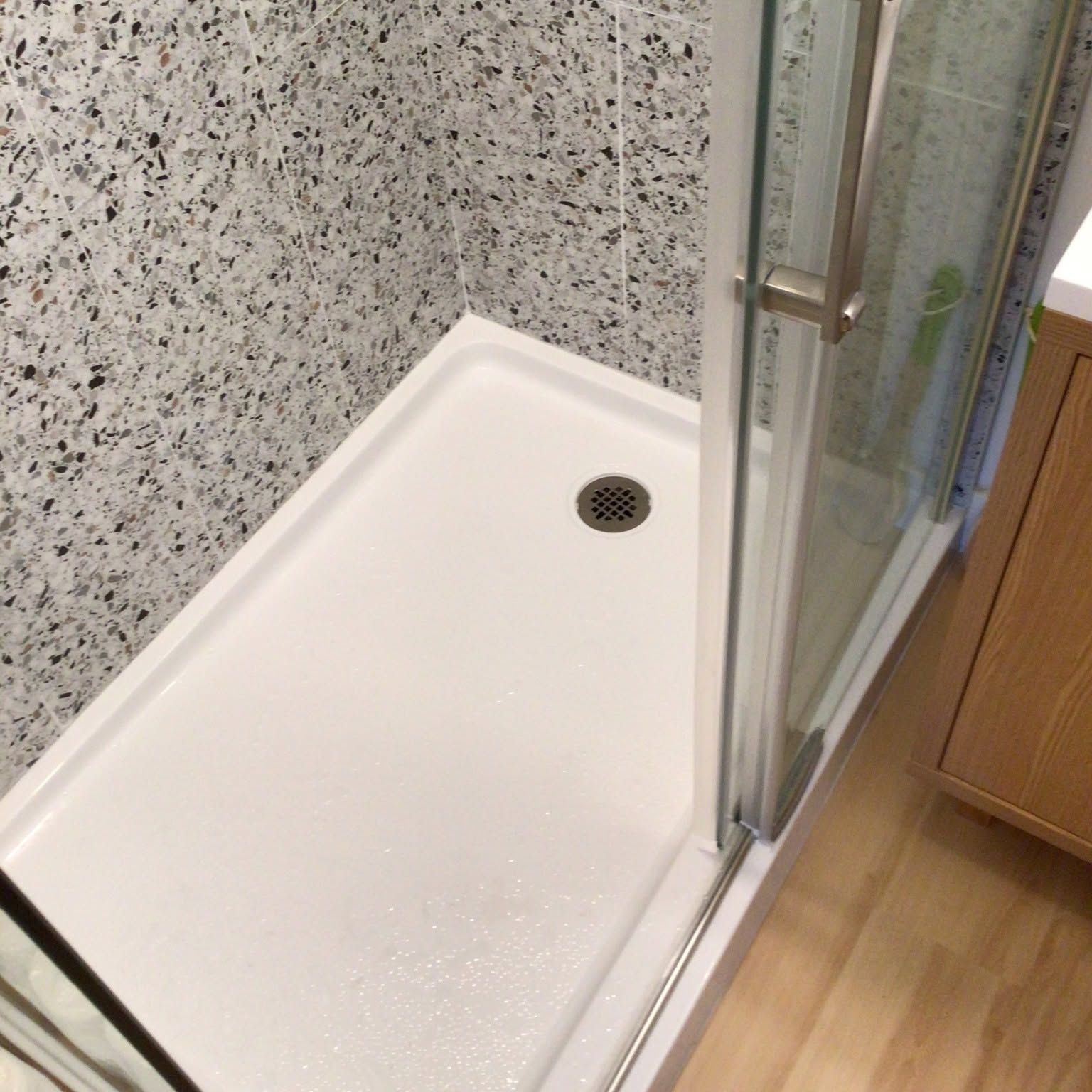 Corner shower stall with white base, glass door, and speckled gray wall tile