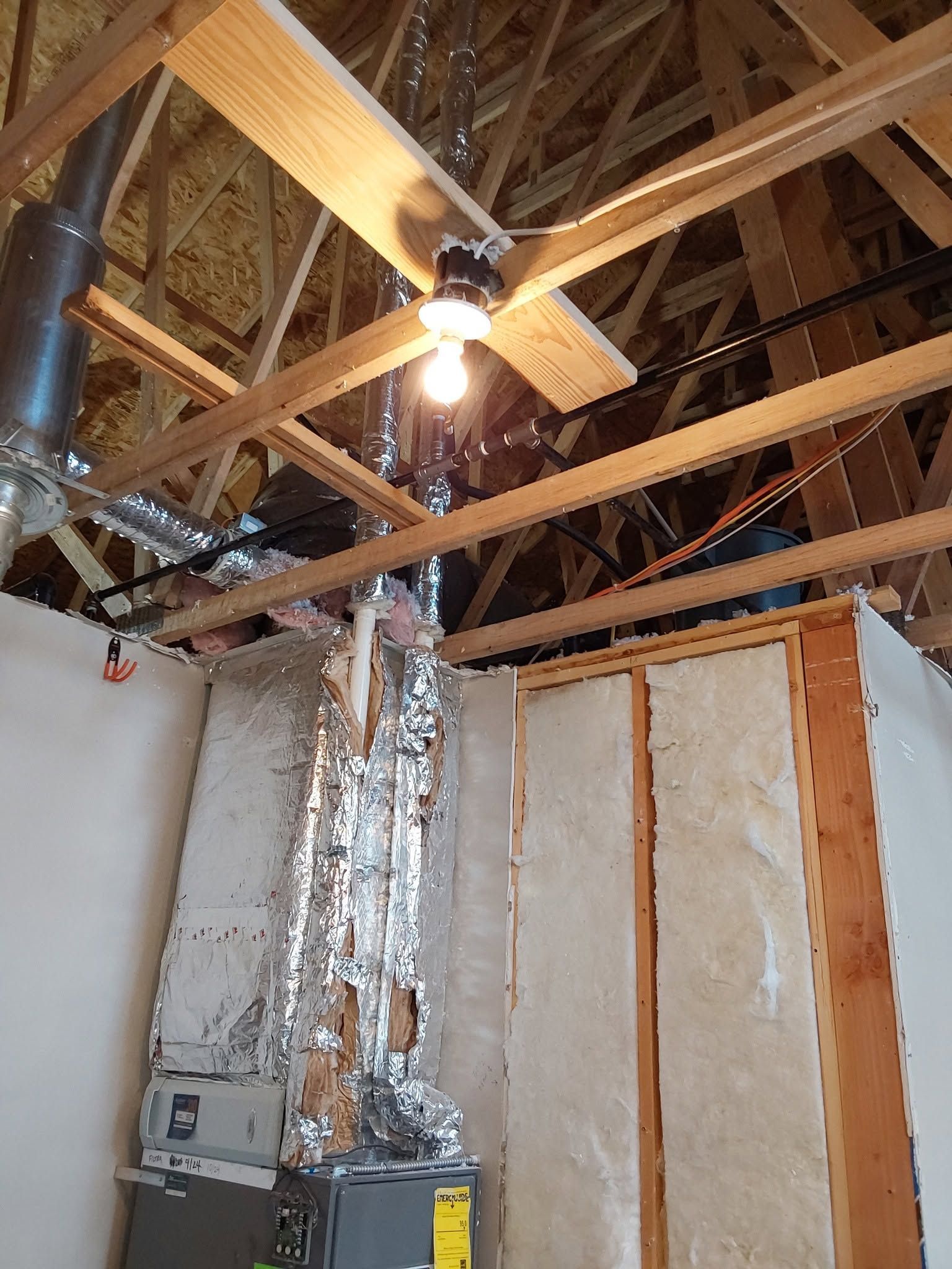 Unfinished room with exposed ceiling beams, hanging light, insulation, and a metal duct against framed walls.