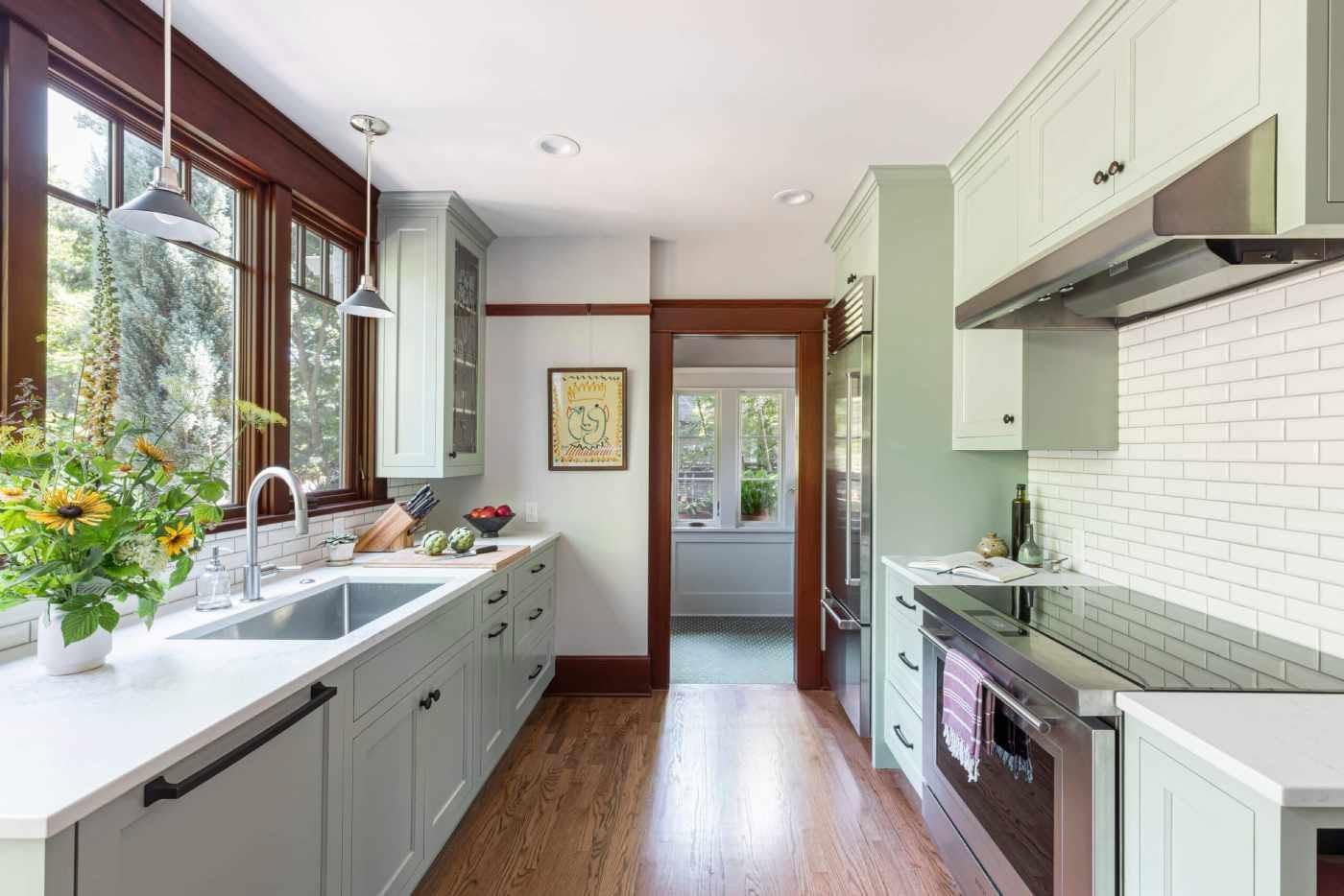 Bright narrow kitchen with white cabinets, gray lower cabinets, wood floor, large window, and green accents