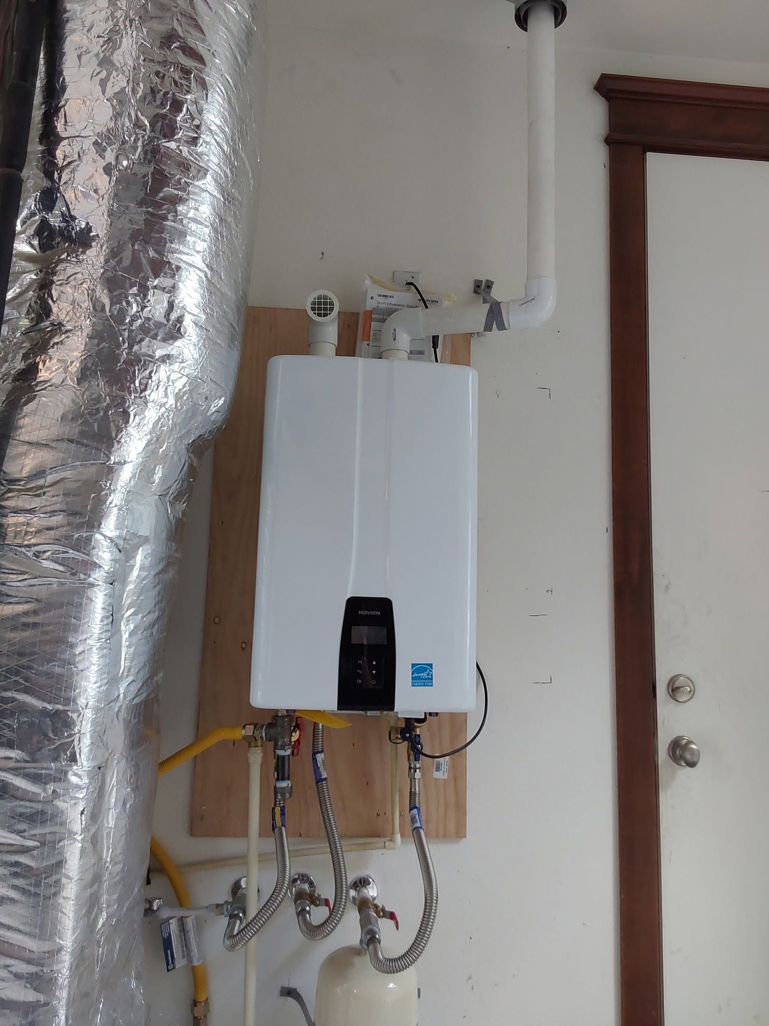 Wall-mounted white tankless water heater with exposed pipes and a silver duct in a utility area