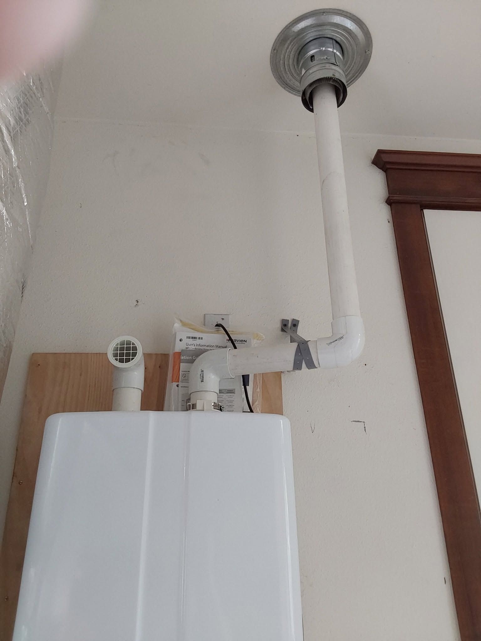Wall-mounted white boiler with exposed white pipe connected to ceiling vent in a room corner