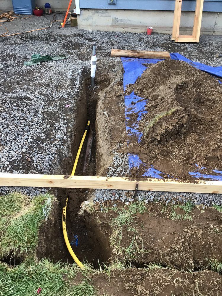 Trench beside a gravel driveway with exposed yellow cable, soil piles, and wooden form boards.