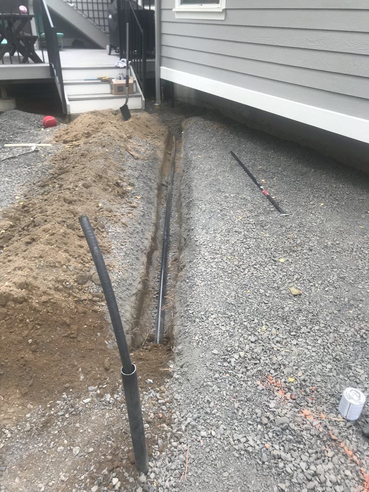 Construction trench beside gray house with exposed pipes and gravel foundation work