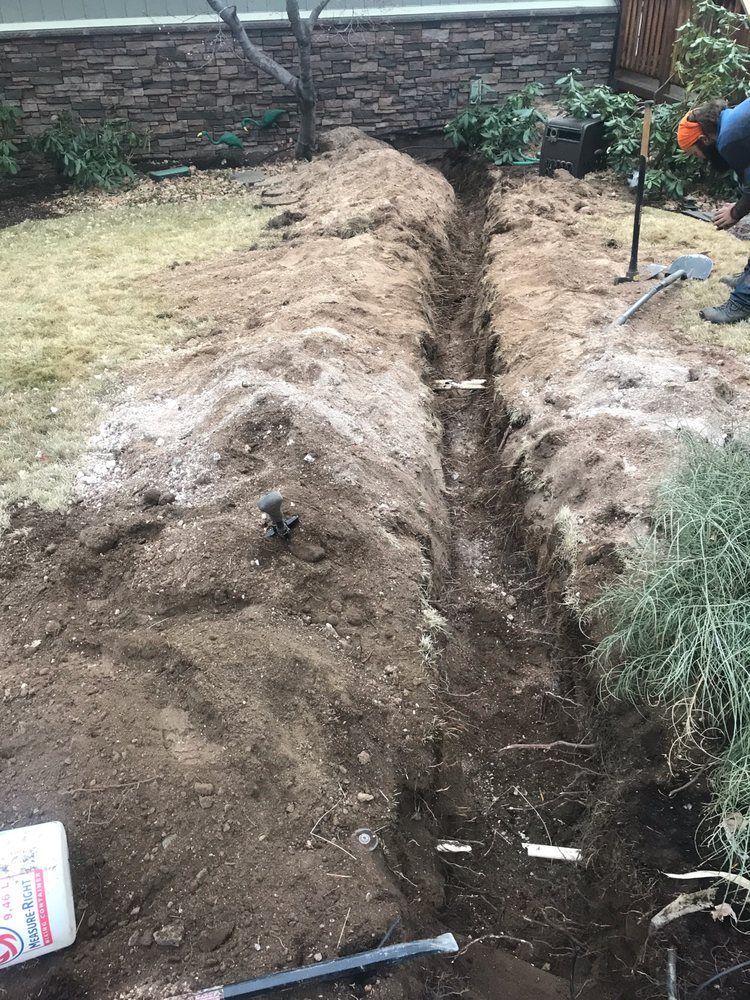 Trench dug in a backyard garden, with piles of soil and a shovel near a brick wall.