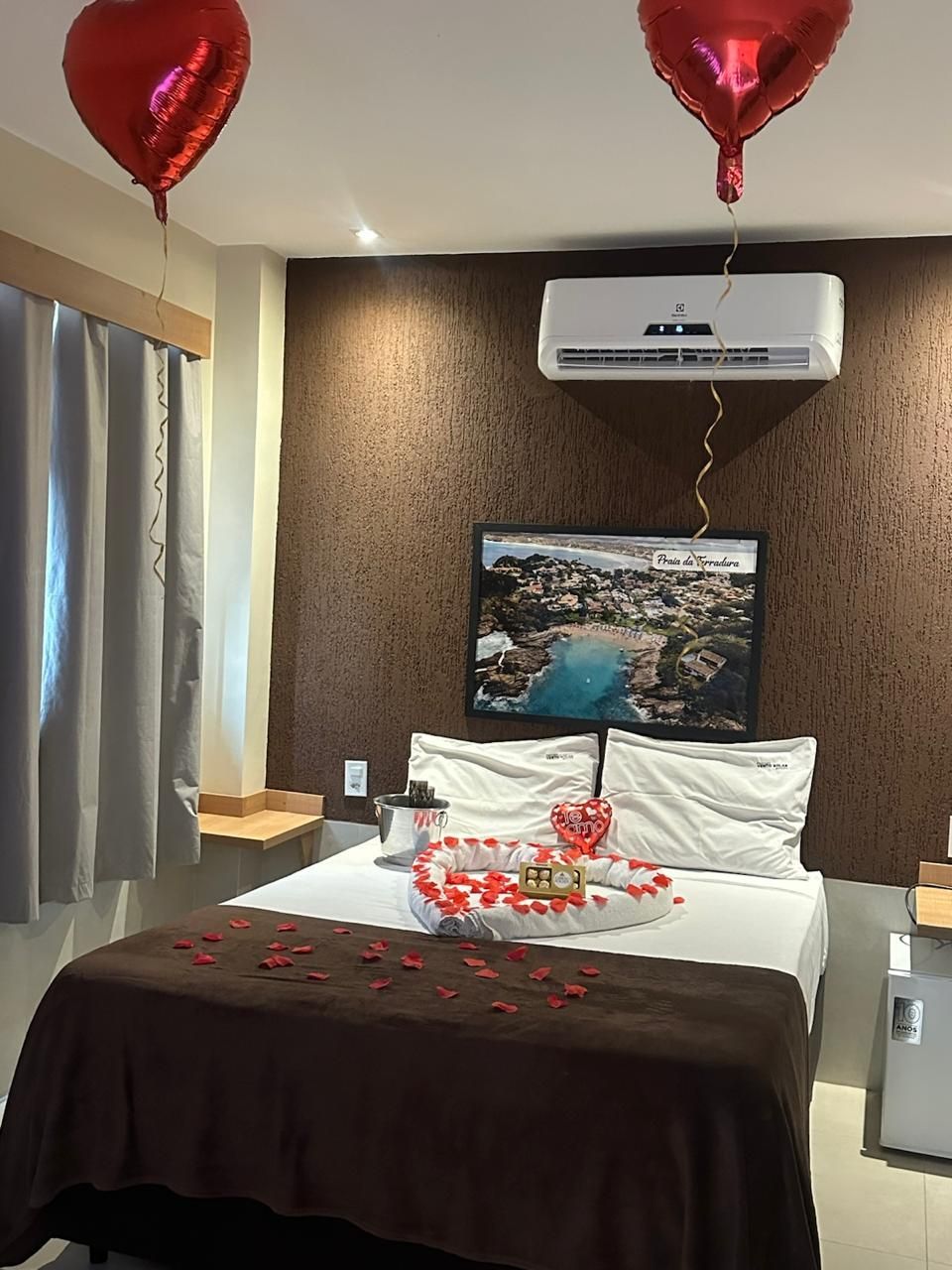 Quarto de hotel com balões em formato de coração, cama decorada com pétalas de rosa e um bolo de aniversário sobre a cama.