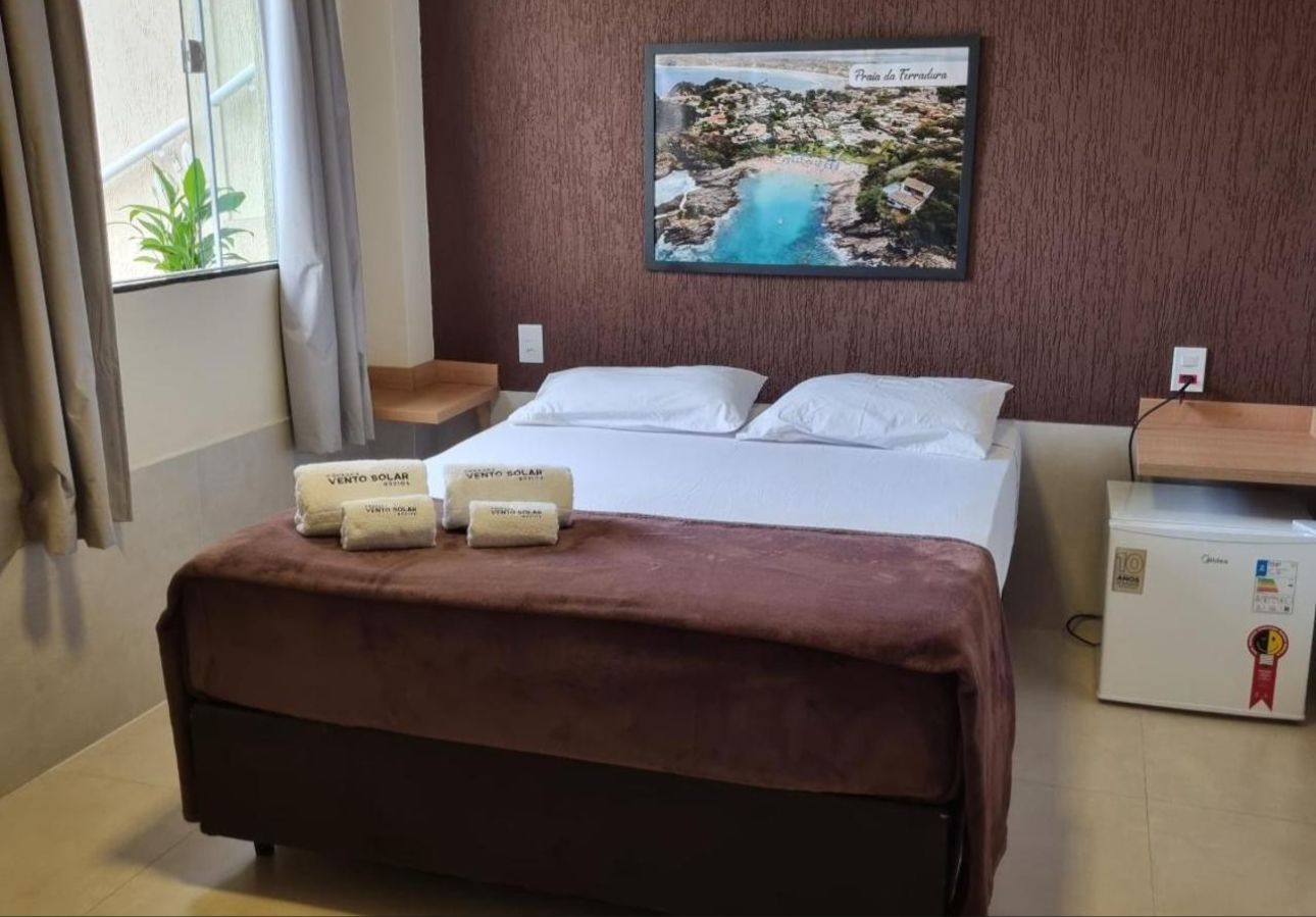 Quarto de hotel com cama de casal, cobertor marrom, escrivaninha pequena e janela com cortinas.