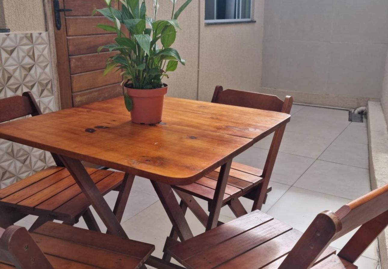 Mesa e cadeiras de madeira para pátio, com um vaso de planta em cima, dispostas em uma varanda com piso de azulejo.