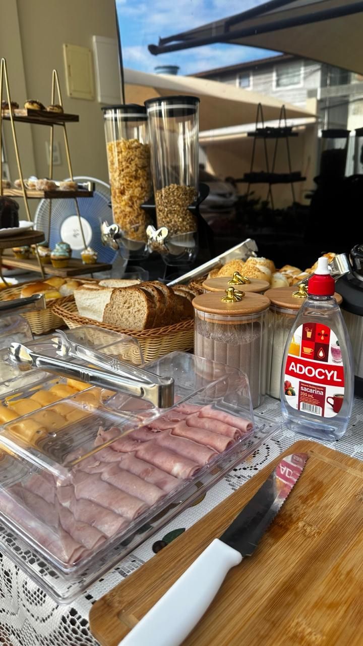 Mesa de bufê com doces, frios, cereais e uma caixa de refrigerante vermelha ao ar livre, perto de uma janela.