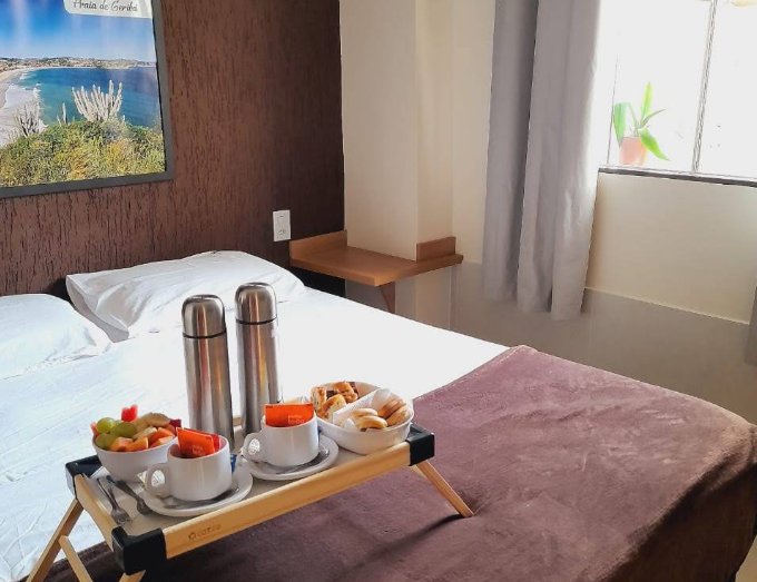 Quarto de hotel com cama arrumada e bandeja de café da manhã sobre as cobertas perto da janela.