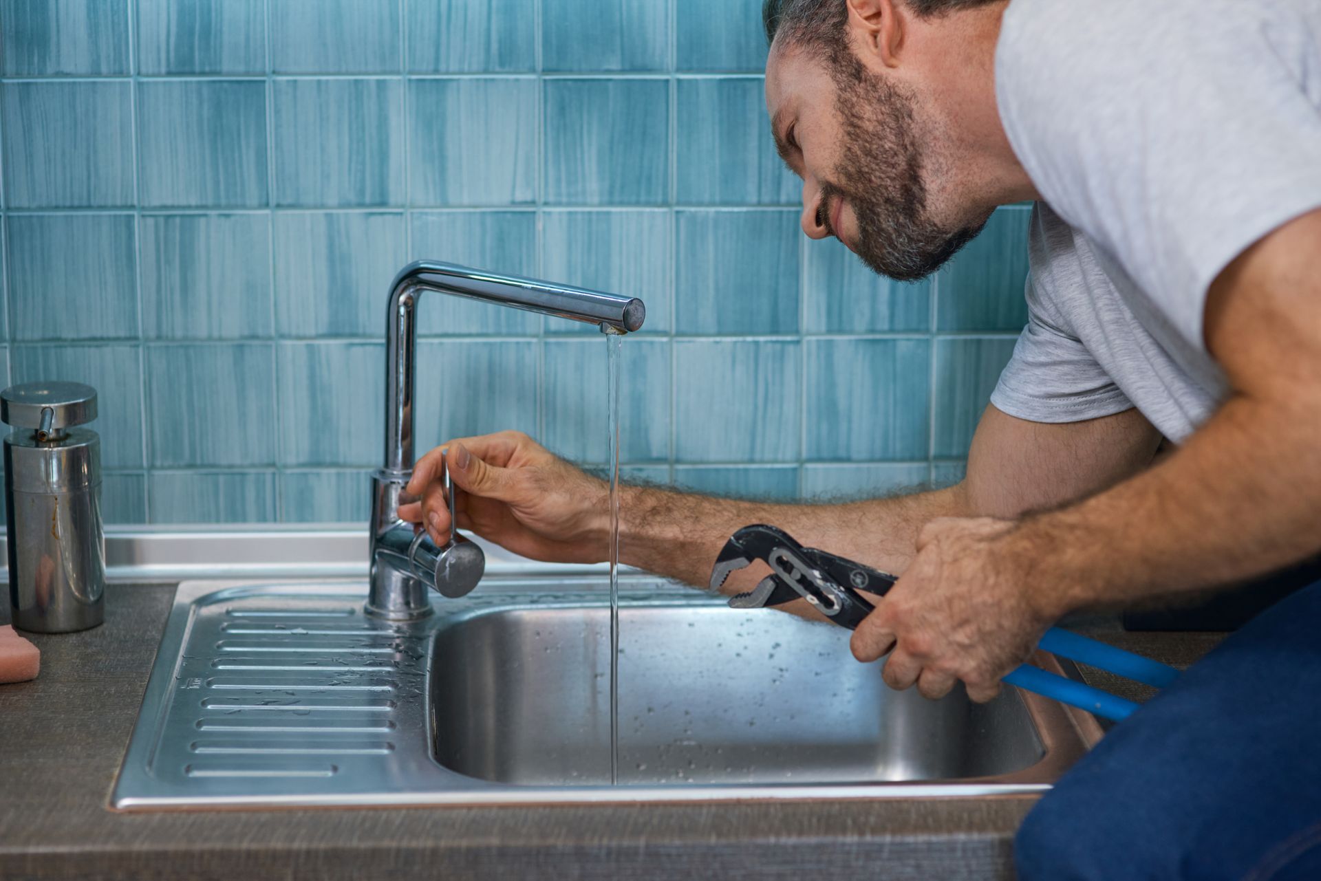 Man repairing a kitchen sink faucet with a wrench beside a stainless steel sink and blue tile wall
