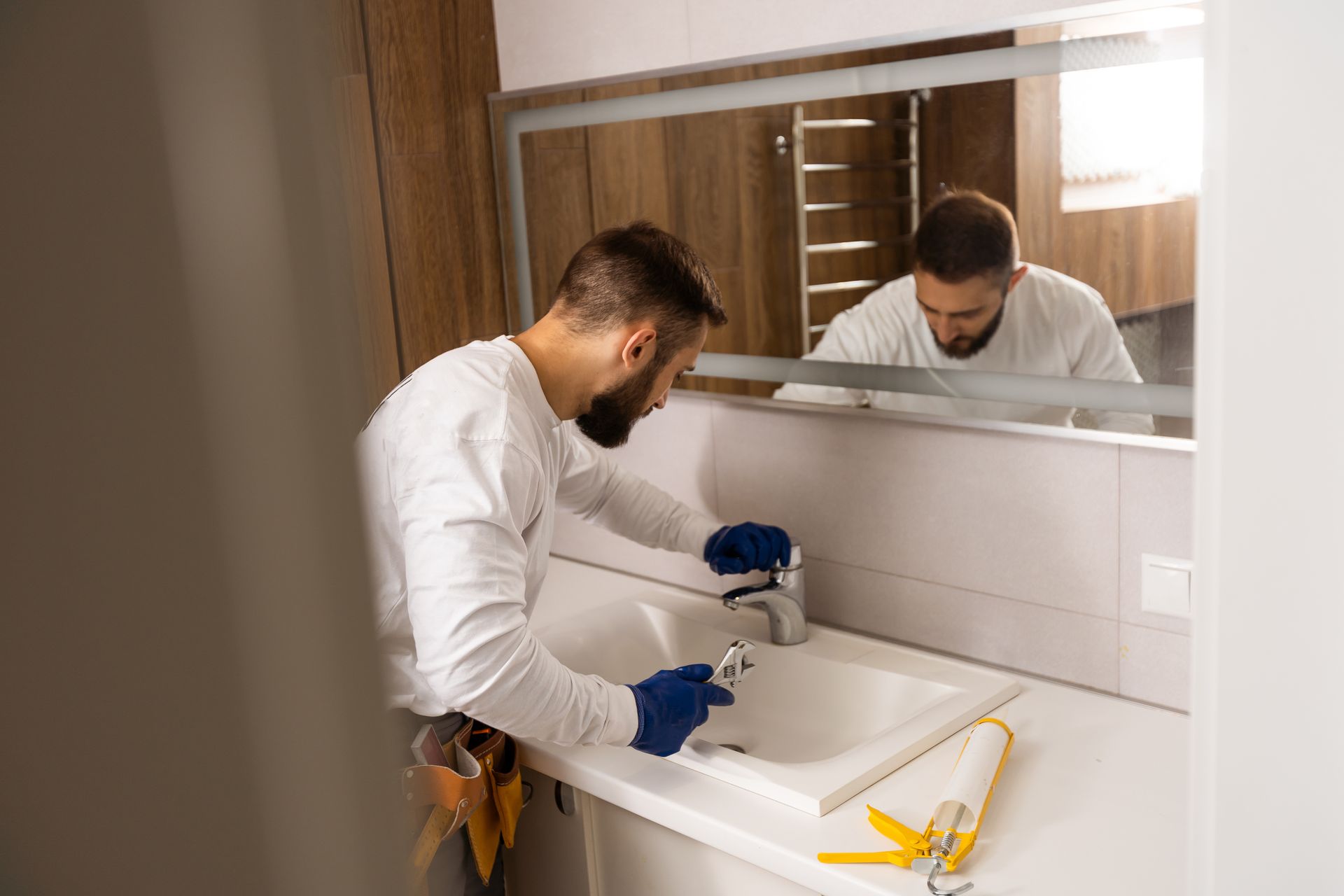 Two plumbers fixing a bathroom sink under a mirror, one using a tool beside a yellow wrench.