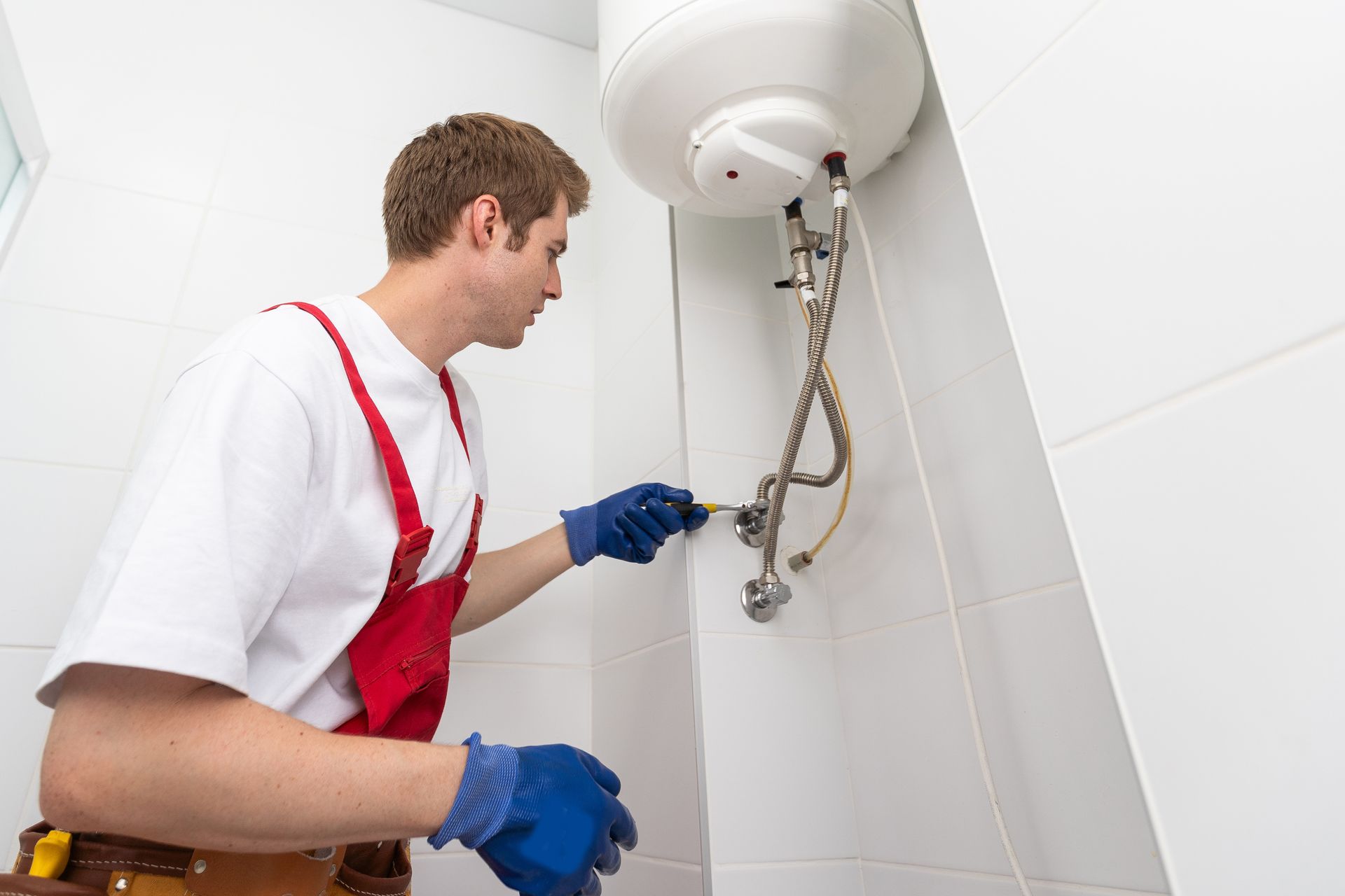 Plumber in red overalls repairs a white bathroom sink pipe under a wall-mounted water heater