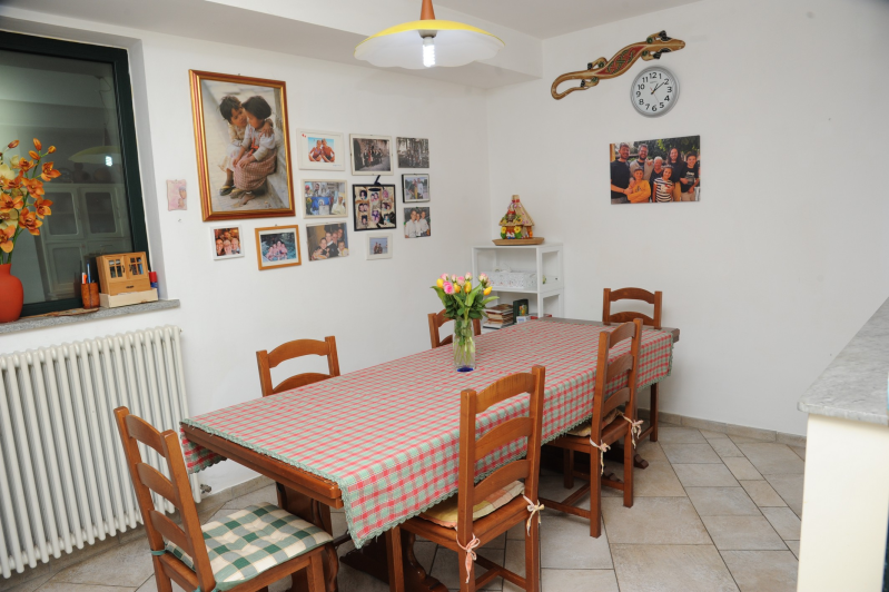 Sala da pranzo con tavolo in legno ricoperto da una tovaglia a quadri rossa e sedie. Foto appese alle pareti bianche.