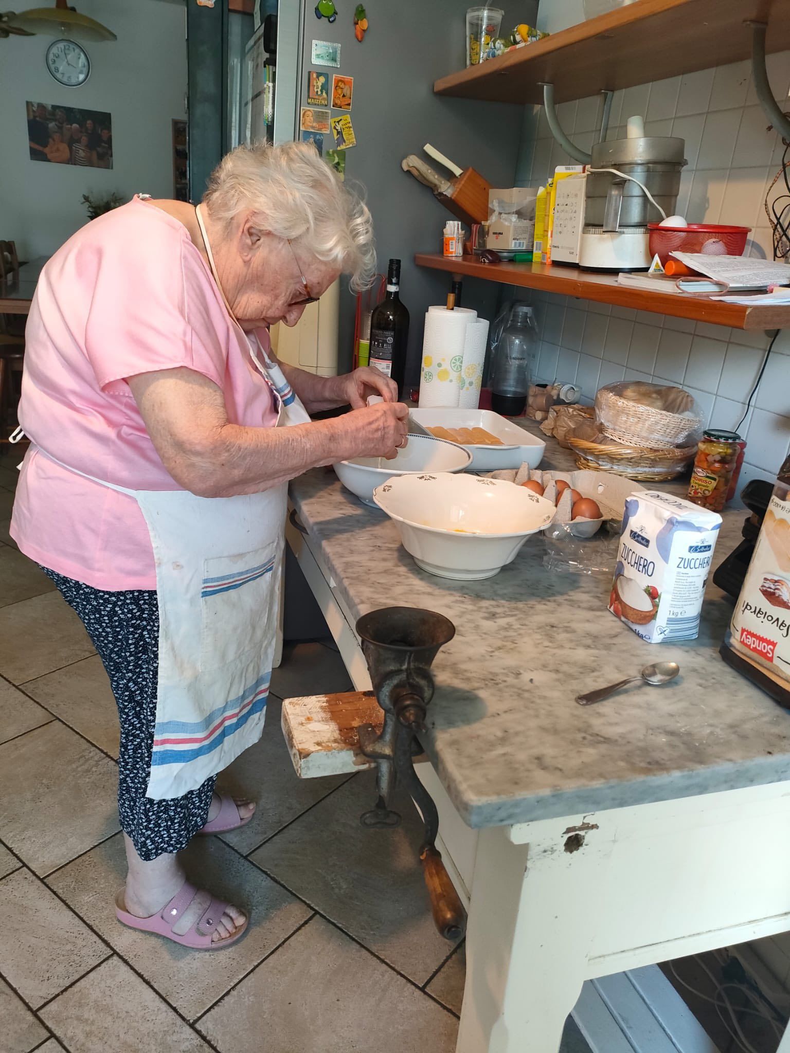Una persona anziana con una camicia rosa e un grembiule cucina e usa un cucchiaio per mescolare gli ingredienti.