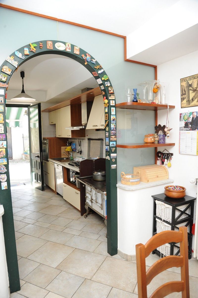 Cucina con porta ad arco, mobili dai colori chiari, pavimento piastrellato, scaffali con oggetti e un tavolo con sedia.