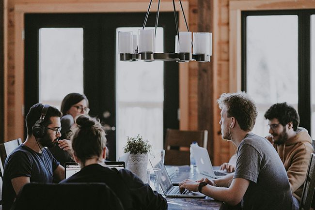 People in a meeting around a table in a bright office with large windows and a hanging light fixture.