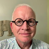 Portrait of a person with round black glasses, smiling in a light green room.